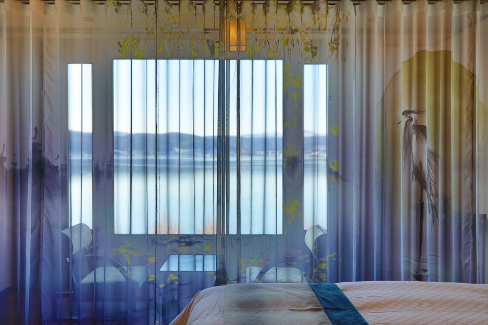 上諏訪温泉「しんゆ」の客室「光神」。水墨画が描かれたオリジナルレースカーテン越しにうっすら覗く諏訪湖を絵画に見立てたしんゆでしか味わえない景色を堪能。